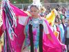 A dancer in a bright pink shawl with fringe, wearing traditional regalia adorned with blue and black accents, performs at an outdoor cultural event.
